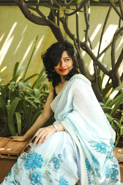 woman in a pastel blue khadi cotton floral printed saree sitting on a bench in a park