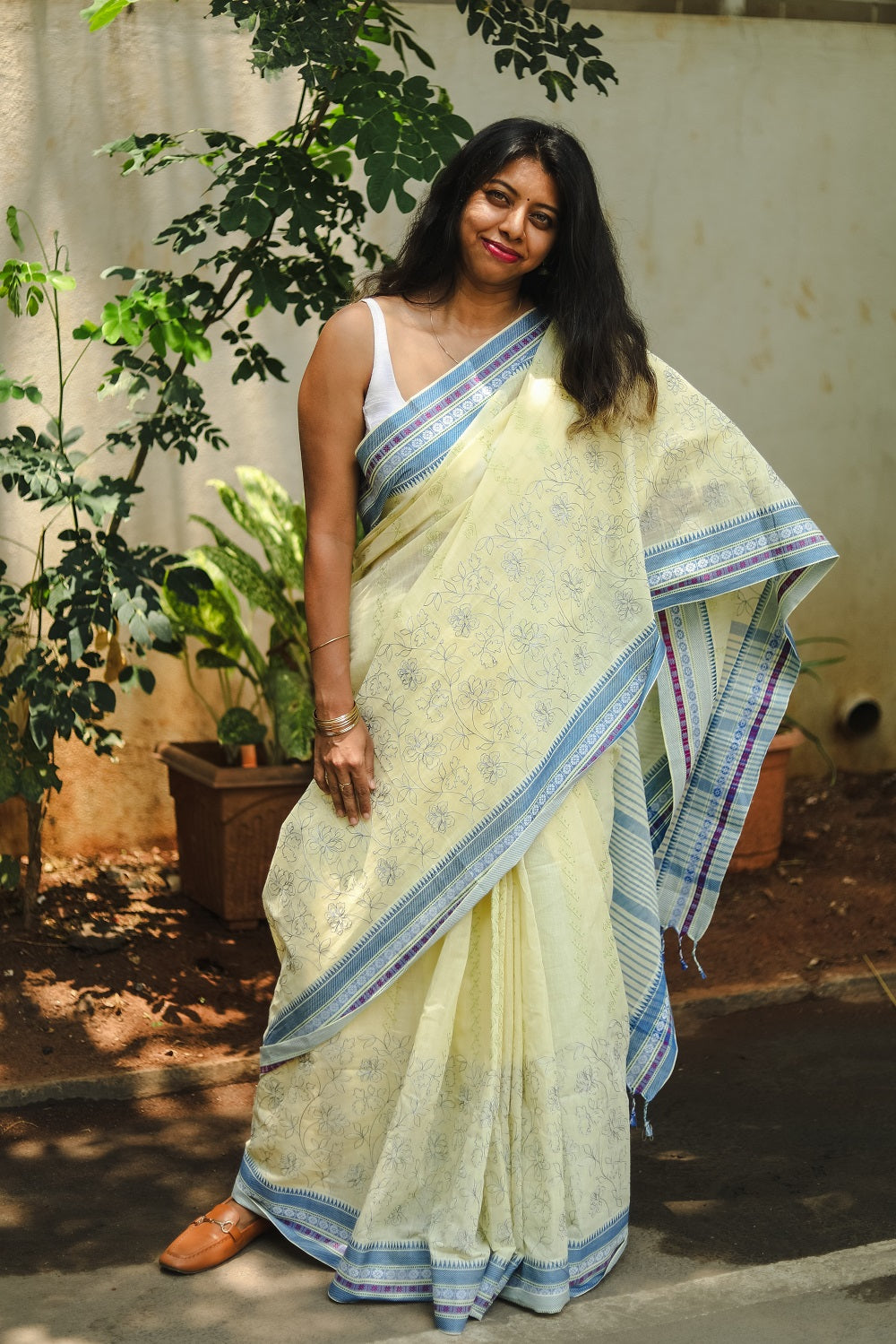 A woman standing in a butter yellow cotton embroidered sari near plants